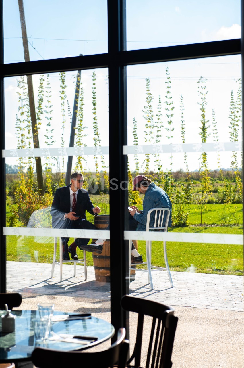 Deux hommes discutent à une table en extérieur, près d'une grande baie vitrée, avec un paysage verdoyant en arrière-plan.