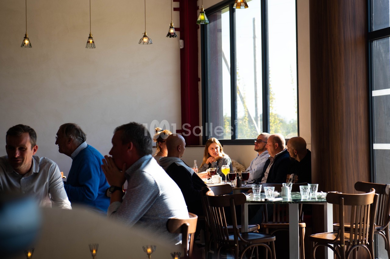 Des personnes assises à des tables dans un restaurant lumineux, discutant et partageant un repas près de grandes fenêtres.