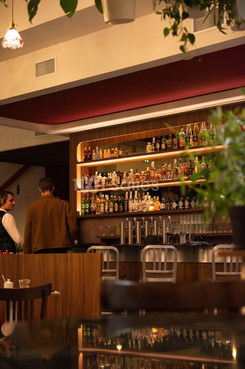 Un bar moderne avec des étagères remplies de bouteilles, deux personnes discutent au comptoir.