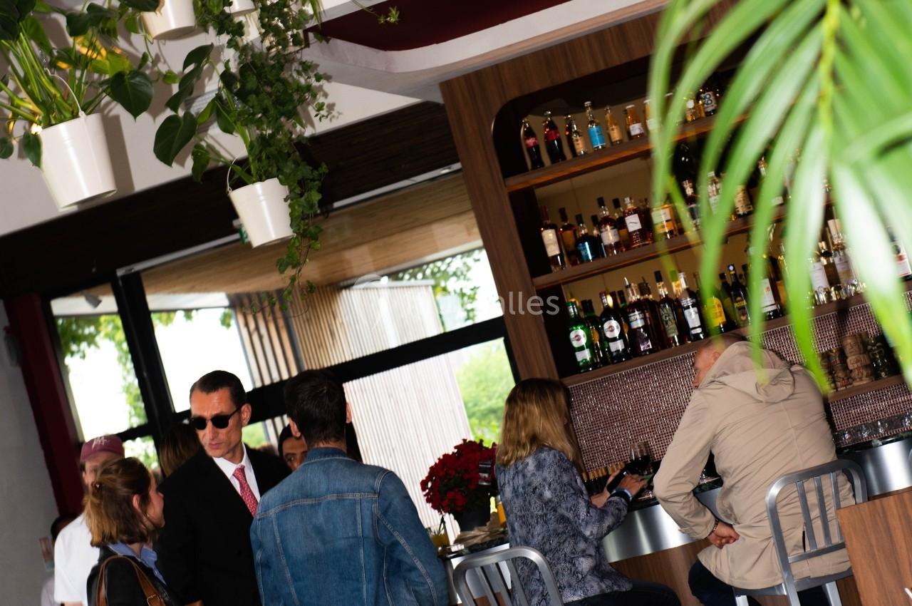 Des personnes discutent et s'installent autour d'un bar dans un espace lumineux avec des plantes suspendues.