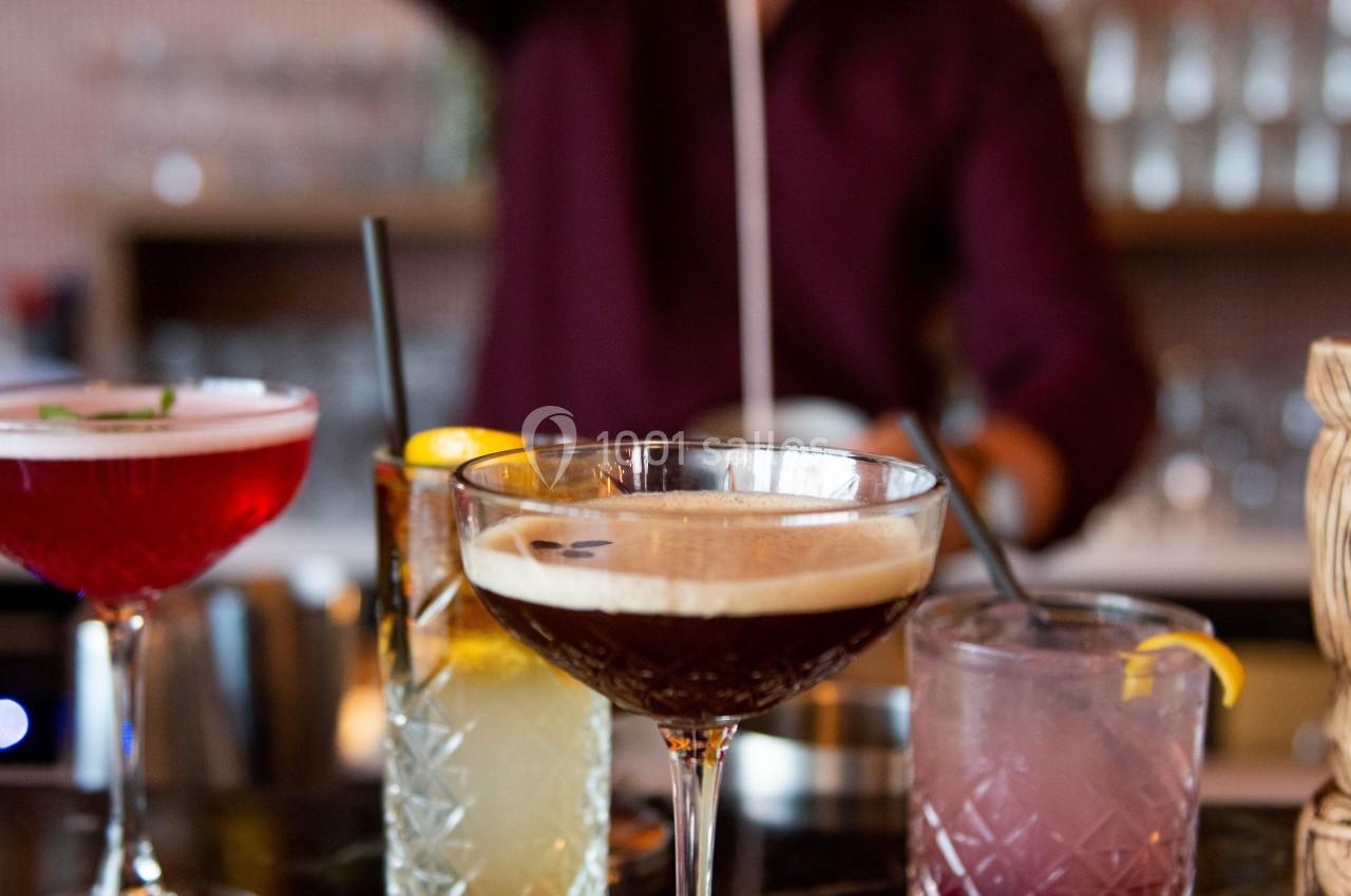Un barman verse un cocktail dans un verre à pied sur un comptoir, entouré d'autres boissons colorées.