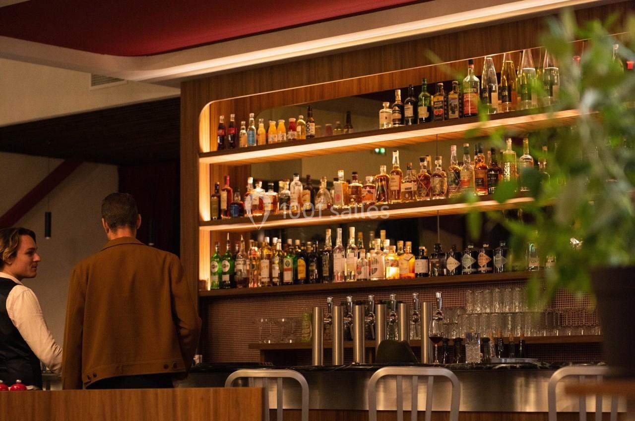 Un bar avec des étagères remplies de bouteilles, un homme debout et un barman discutant dans une ambiance chaleureuse.