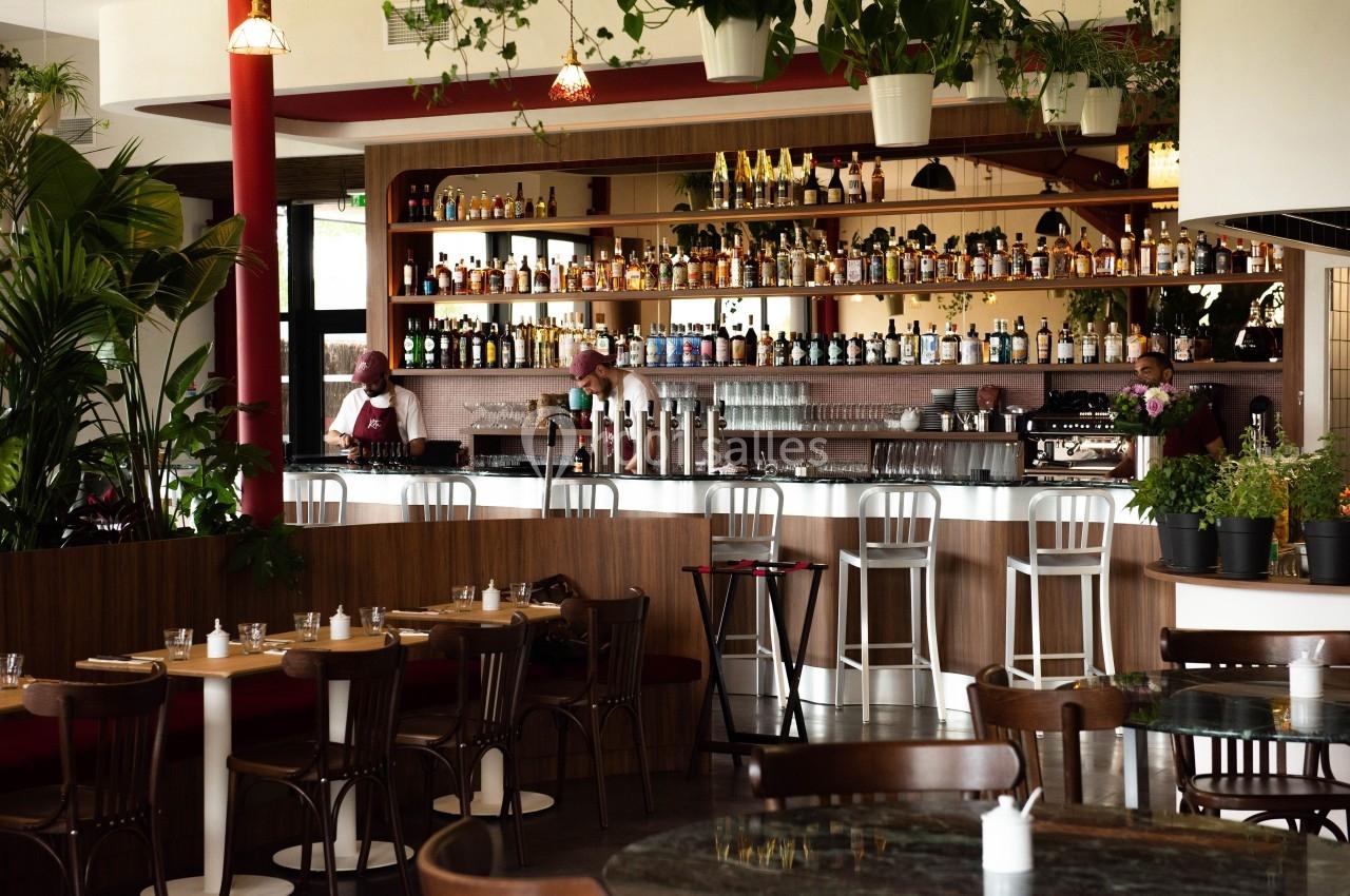 Bar moderne avec étagères remplies de bouteilles, comptoir éclairé, chaises hautes et tables entourées de plantes.