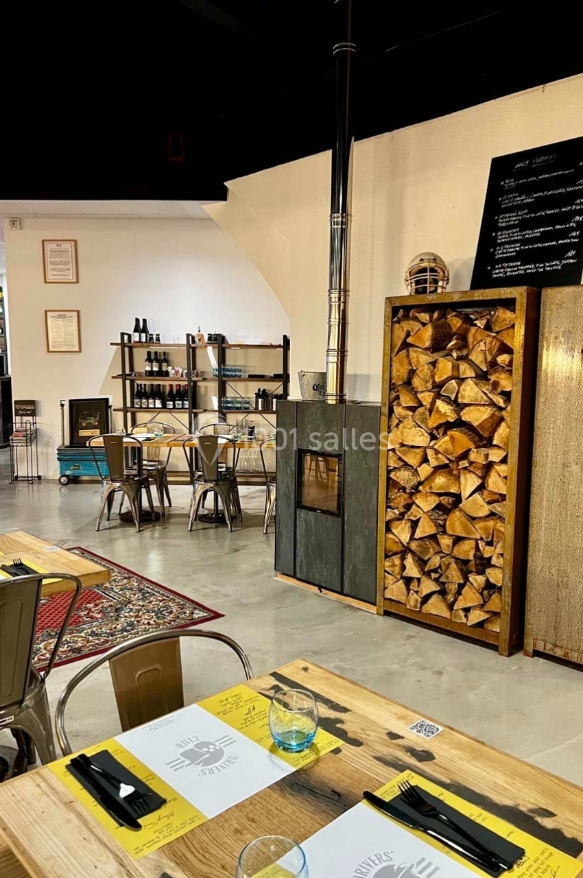 Salle de restaurant chaleureuse avec tables en bois, poêle à bois, étagères de bouteilles et ambiance conviviale.