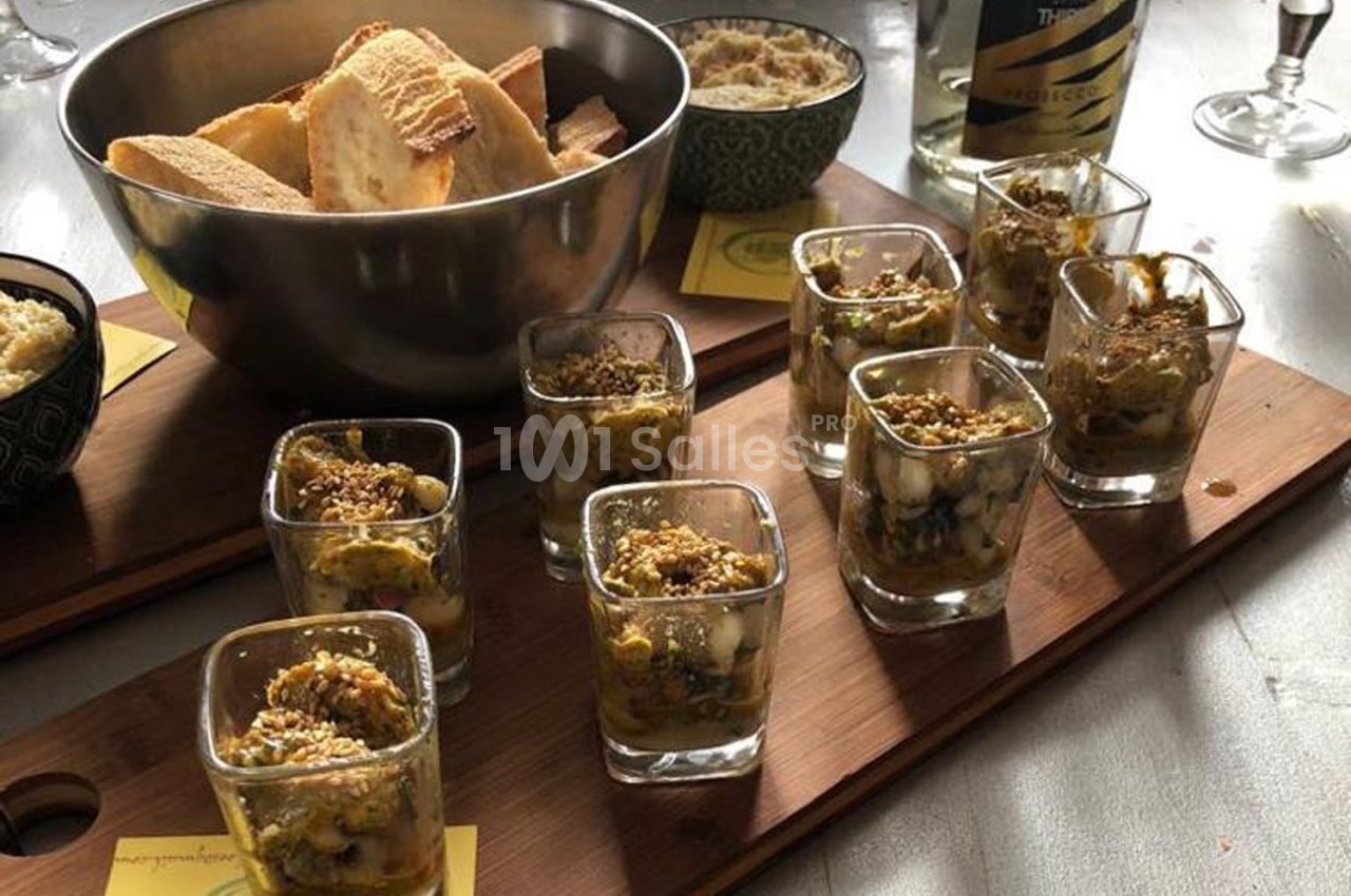 Plateau en bois avec verrines garnies, bol de pain tranché et bouteille de vin mousseux en arrière-plan.