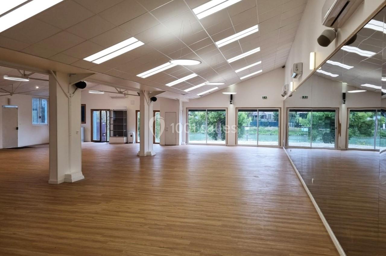Salle spacieuse avec parquet en bois clair, murs blancs, grandes baies vitrées et miroirs sur un côté.