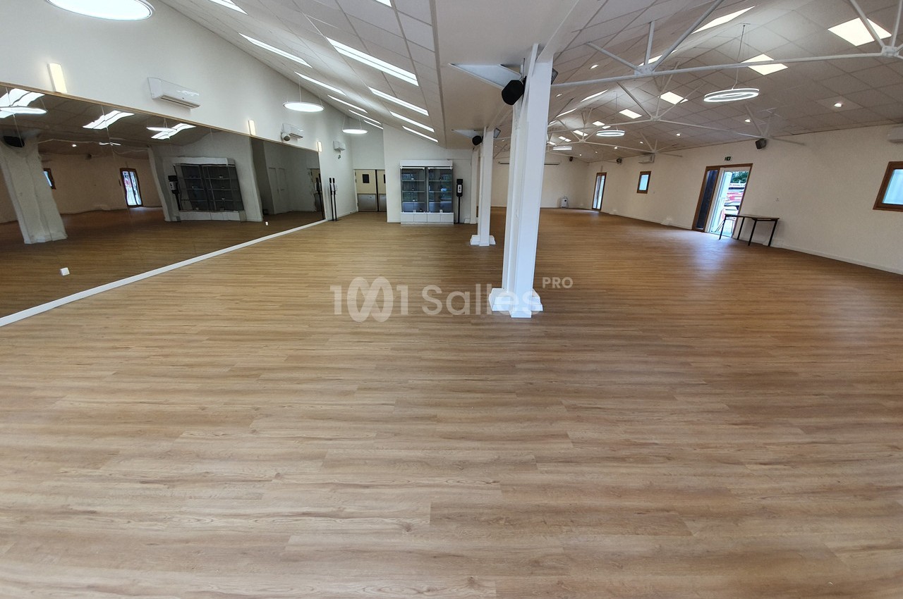 Salle spacieuse avec parquet en bois clair, murs blancs, miroirs et éclairage naturel provenant de fenêtres.