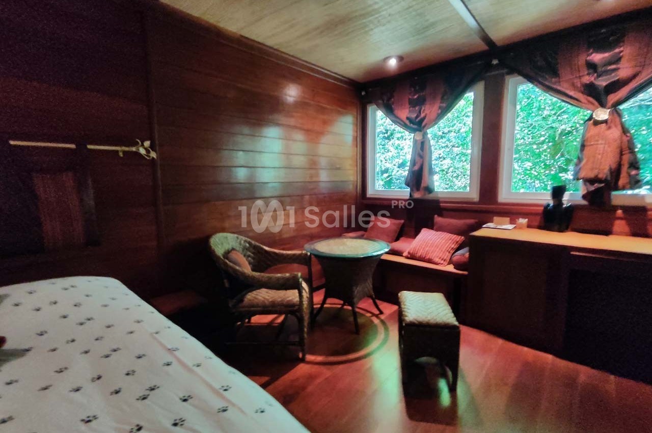 Chambre en bois avec lit, fauteuils en osier, table ronde, bureau et fenêtres donnant sur un paysage verdoyant.
