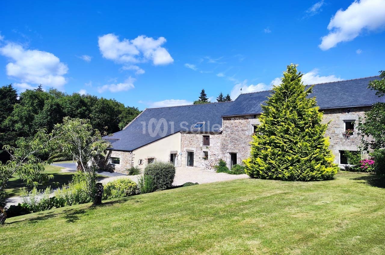 Bâtisse en pierre avec toit en ardoise entourée de verdure et d'un jardin sous un ciel bleu.
