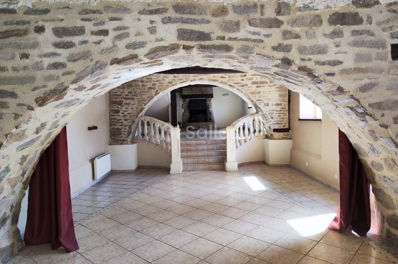 Salle voûtée en pierre avec sol carrelé, escaliers menant à une cheminée et rideaux rouges encadrant les ouvertures.