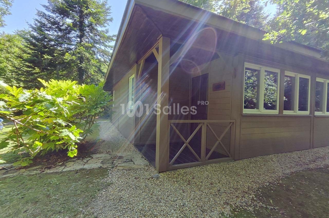 Chalet en bois entouré de végétation, avec une allée de gravier et des rayons de soleil traversant les arbres.