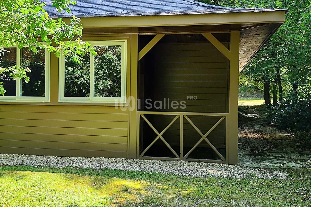 Cabane en bois avec fenêtres et porche ouvert, entourée de verdure et éclairée par la lumière naturelle.