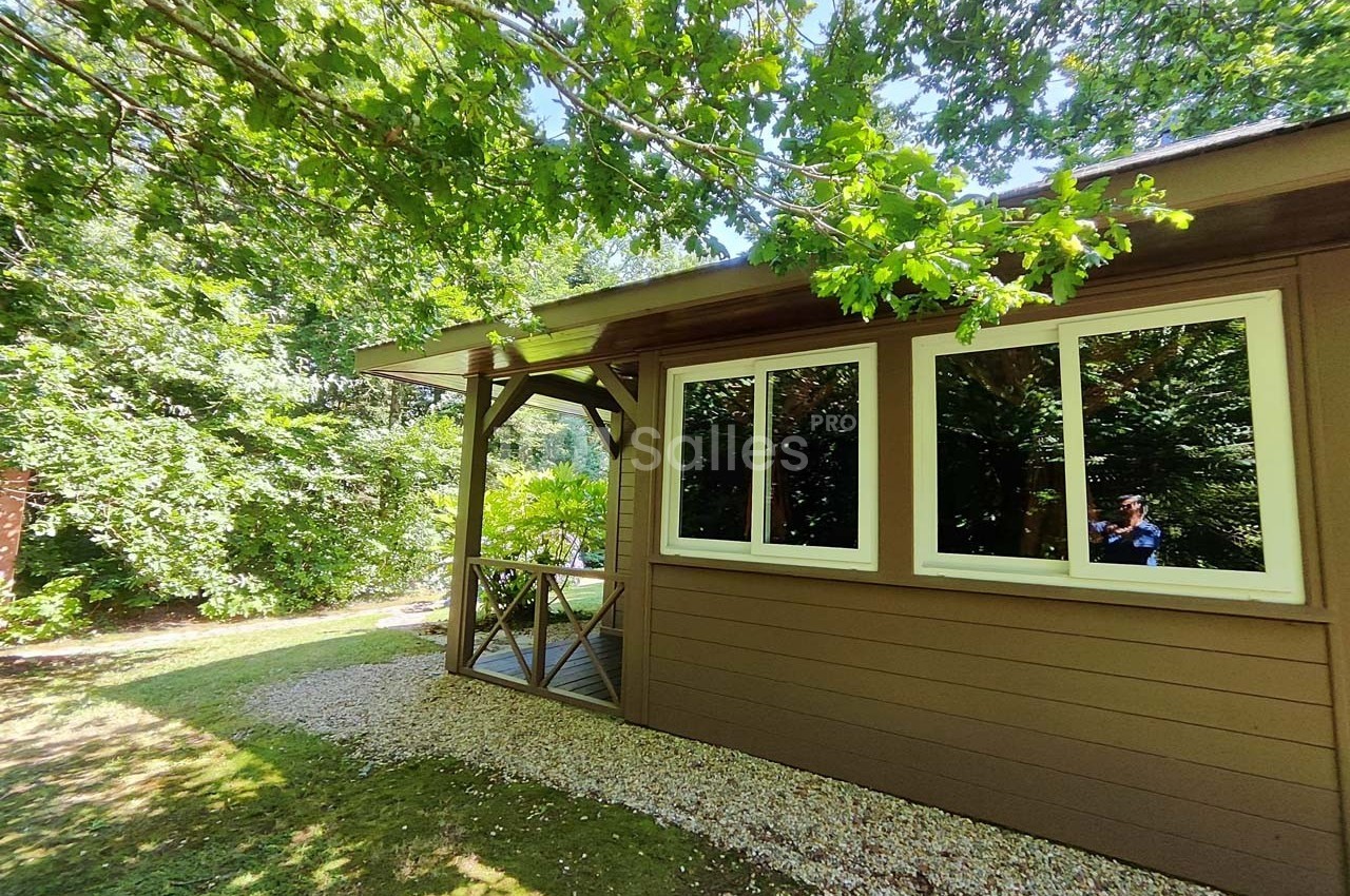 Maison en bois avec terrasse couverte, entourée de verdure et éclairée par la lumière naturelle.