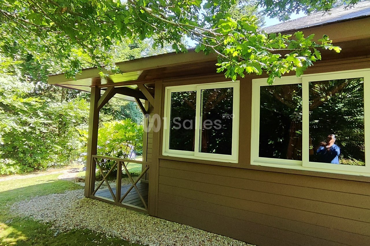 Maison en bois avec terrasse couverte, entourée de verdure et éclairée par la lumière naturelle.