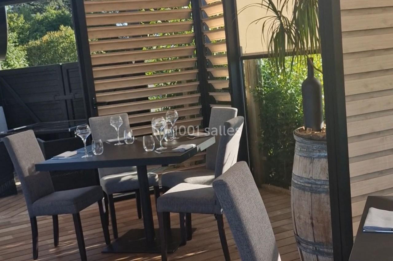 Terrasse en bois avec table dressée, chaises grises, parois en lames de bois et plante en pot sur un tonneau.