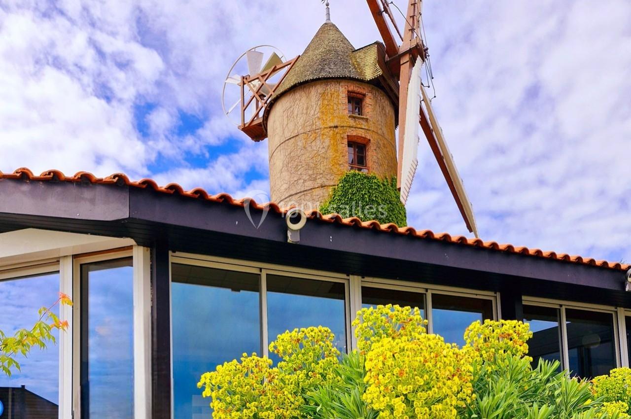 Vue d'un moulin à vent en pierre avec des ailes, partiellement entouré de végétation et surplombant un bâtiment moderne.