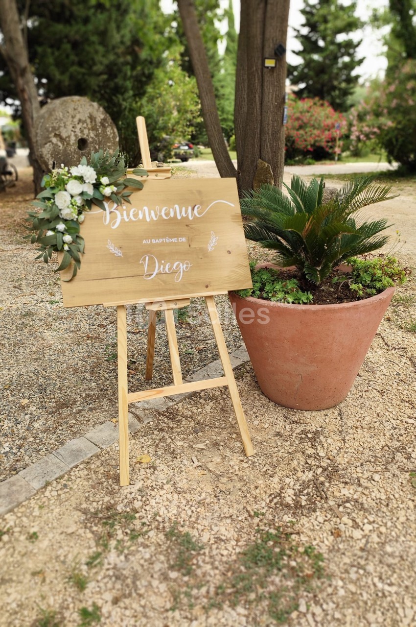 Panneau en bois sur chevalet avec fleurs, inscrit ’Bienvenue au baptême de Diego’, placé dans un jardin.