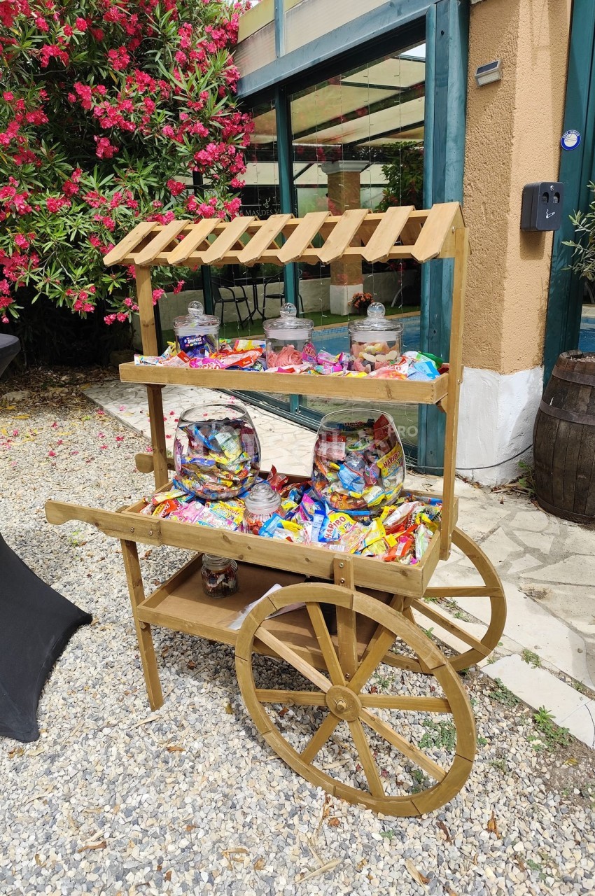 Chariot en bois rempli de bonbons colorés dans des bocaux et sachets, placé sur un sol en gravier près d'une végétation…
