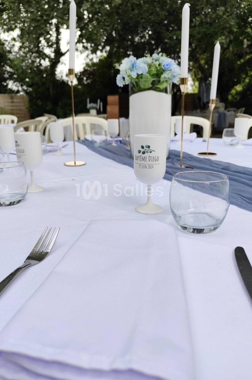 Table dressée en extérieur avec nappes blanches, verres, couverts, bougies et centre de table floral.