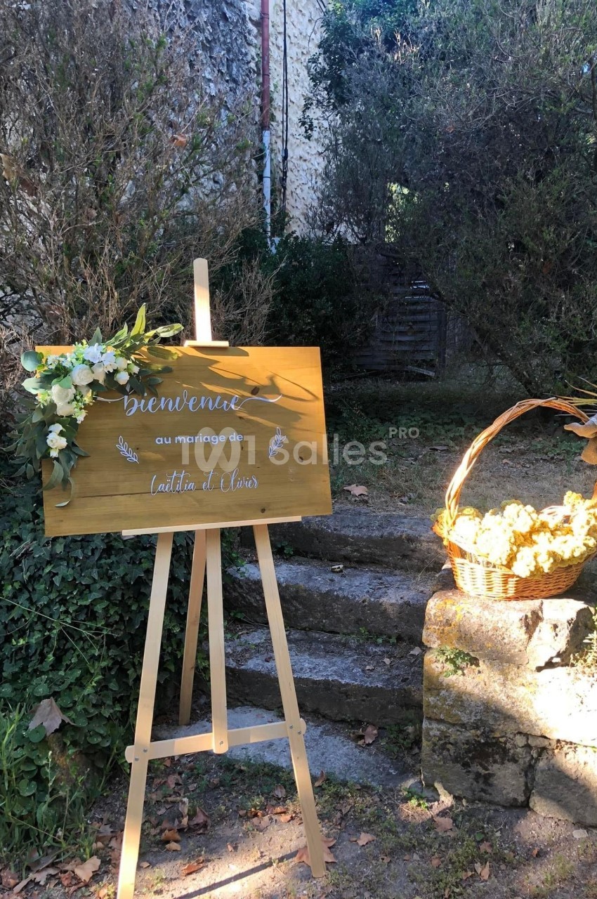 Panneau en bois décoré de fleurs blanches, posé sur un chevalet, à côté d'un panier de fleurs jaunes dans un jardin.