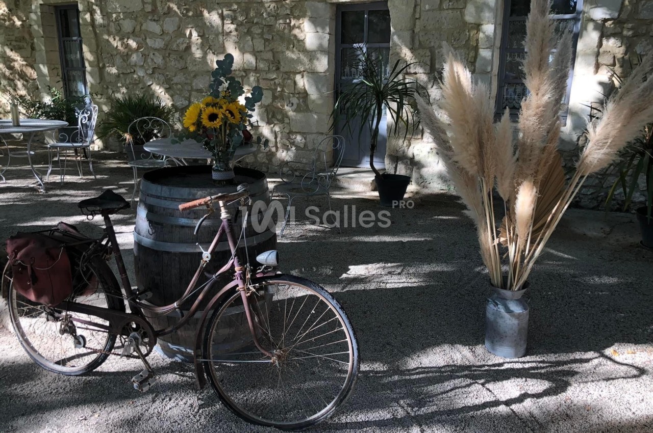 Un vélo ancien posé près d'un tonneau décoré de fleurs, devant un mur en pierre et des plantes en pot.