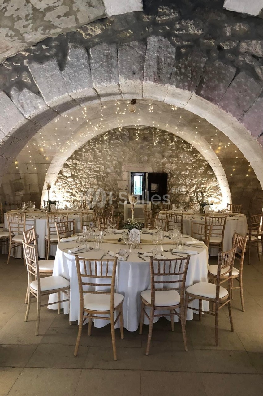 Salle voûtée en pierre décorée de guirlandes lumineuses, avec des tables rondes dressées pour un événement.
