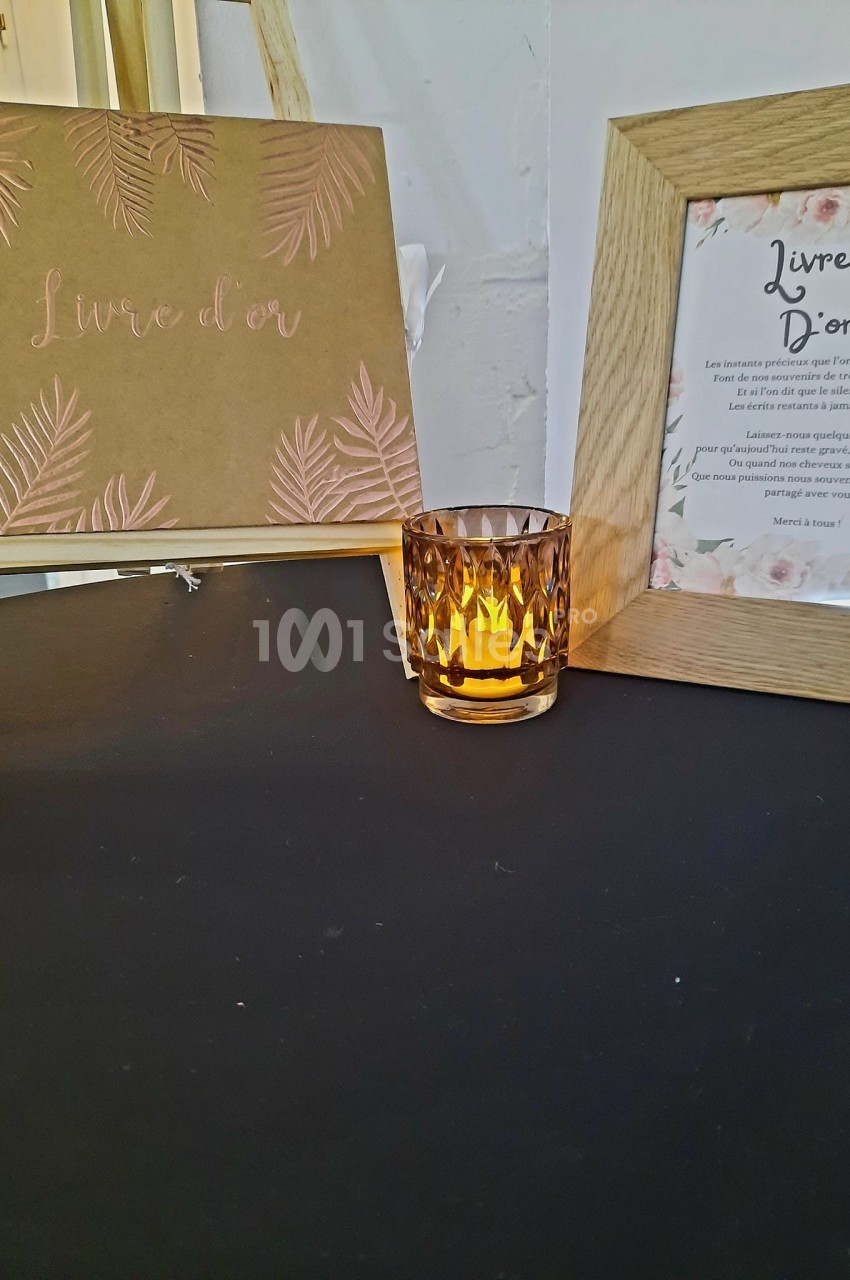 Livre d'or décoratif sur un chevalet, bougie ambrée allumée et cadre en bois sur une table noire.