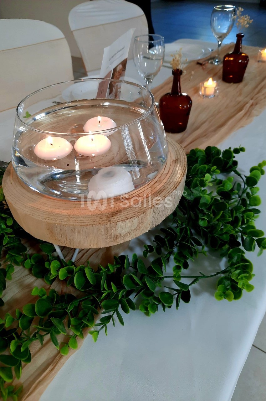 Bougies flottantes dans un vase en verre sur support en bois, entouré de feuillage et décorations de table.
