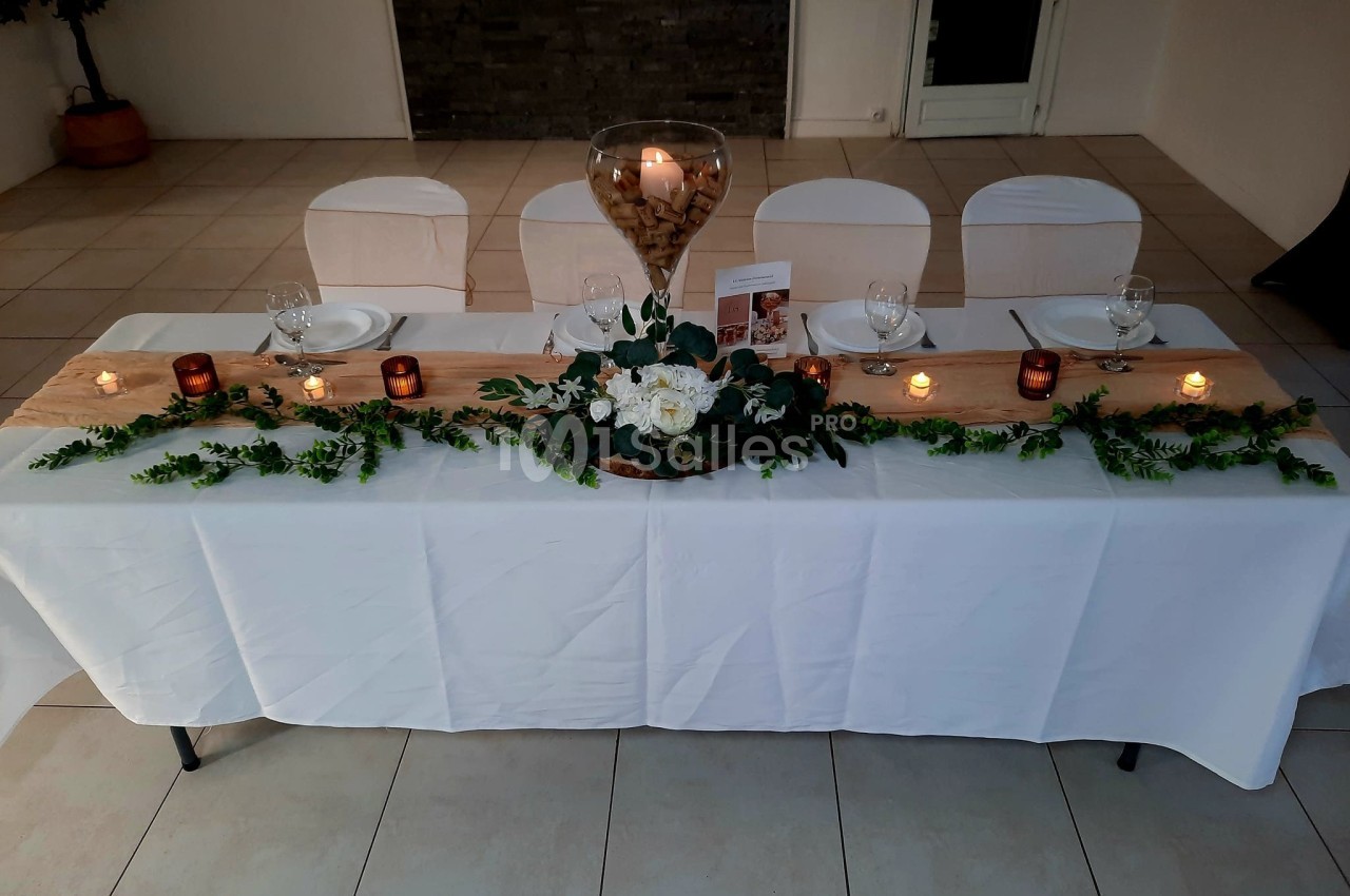 Table décorée pour un événement, ornée de bougies, fleurs blanches, feuillage et un vase central rempli de bouchons en liège.