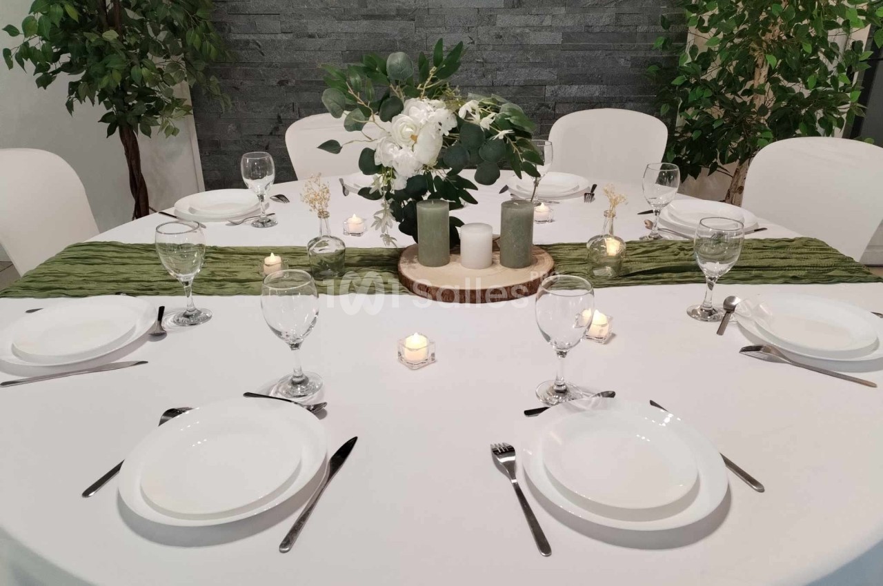 Table ronde élégamment dressée avec nappe blanche, chemin de table vert, bougies, vaisselle et centre de table floral.