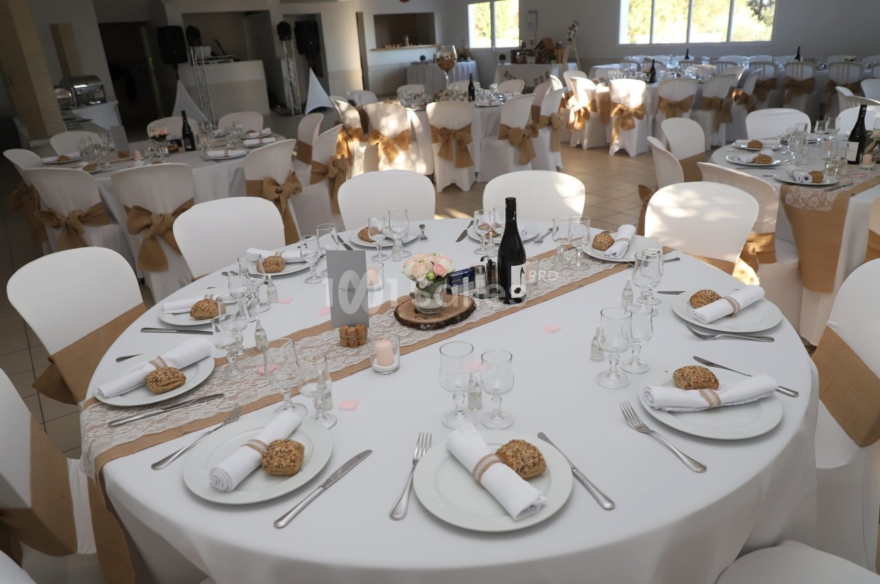 Salle de réception avec tables rondes dressées, nappes blanches, chemins de table en dentelle et décoration élégante.