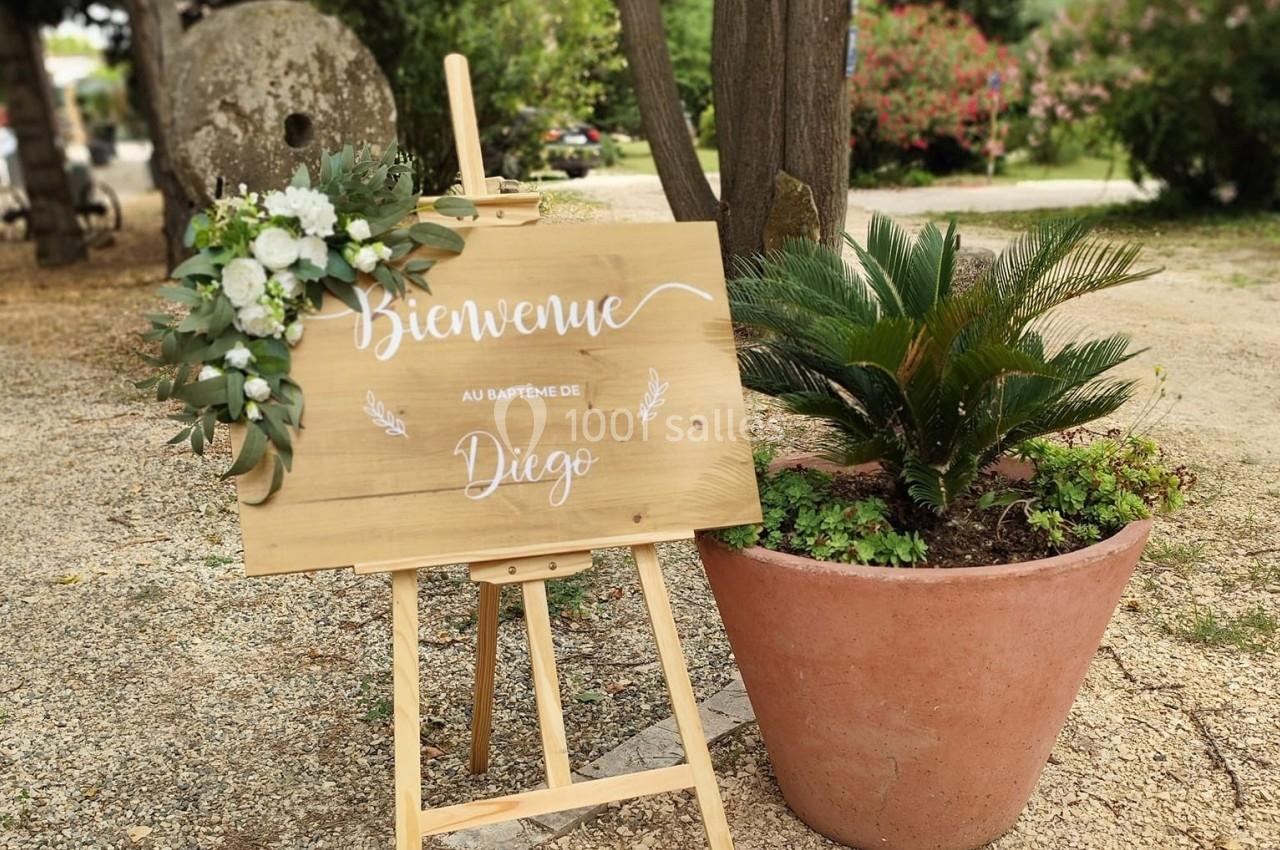 Panneau en bois décoré de fleurs, posé sur un chevalet, indiquant ’Bienvenue au baptême de Diego’, à côté d'une plante en…