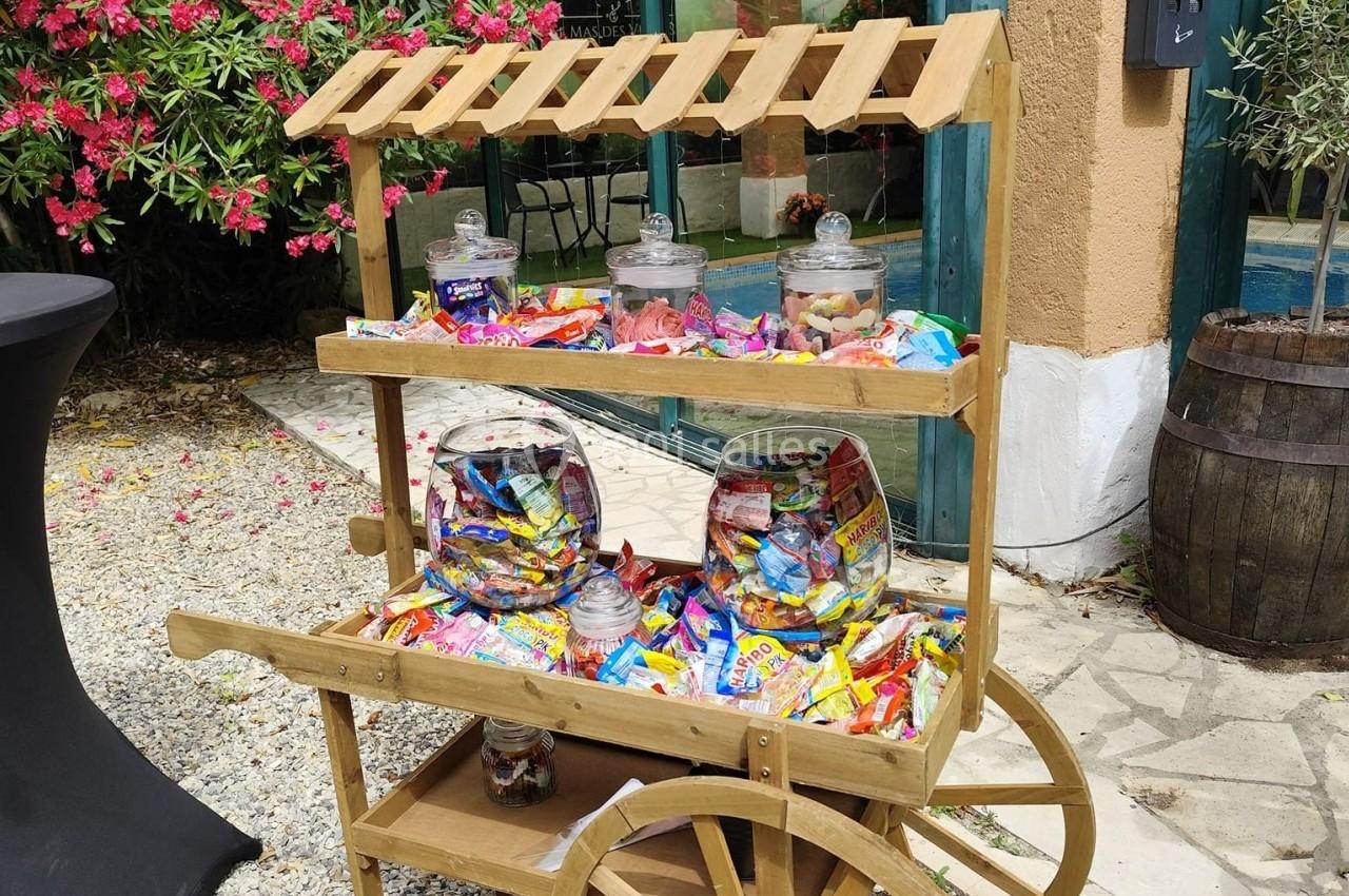 Chariot en bois rempli de bonbons variés dans des bocaux et sachets, placé en extérieur devant un bâtiment.
