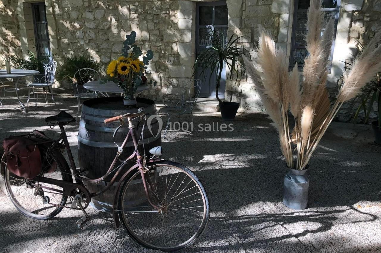 Vélo ancien posé près d'un tonneau décoré de fleurs, sur une terrasse ensoleillée avec des plantes et des meubles en fer…