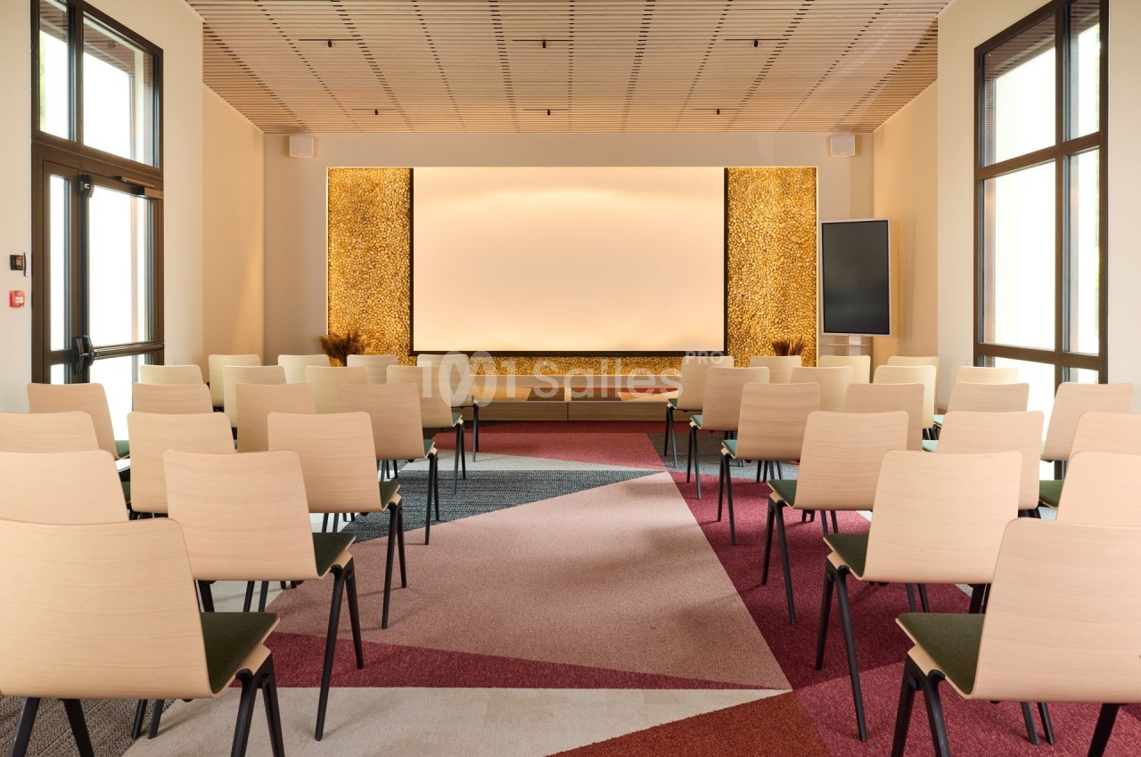 Salle de conférence lumineuse avec des rangées de chaises alignées face à un écran blanc et des murs décoratifs.