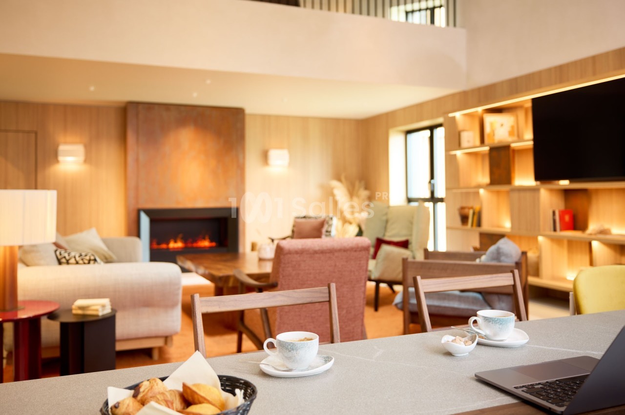 Espace de vie lumineux avec cheminée, canapé, fauteuils, étagères et table avec café et viennoiseries au premier plan.