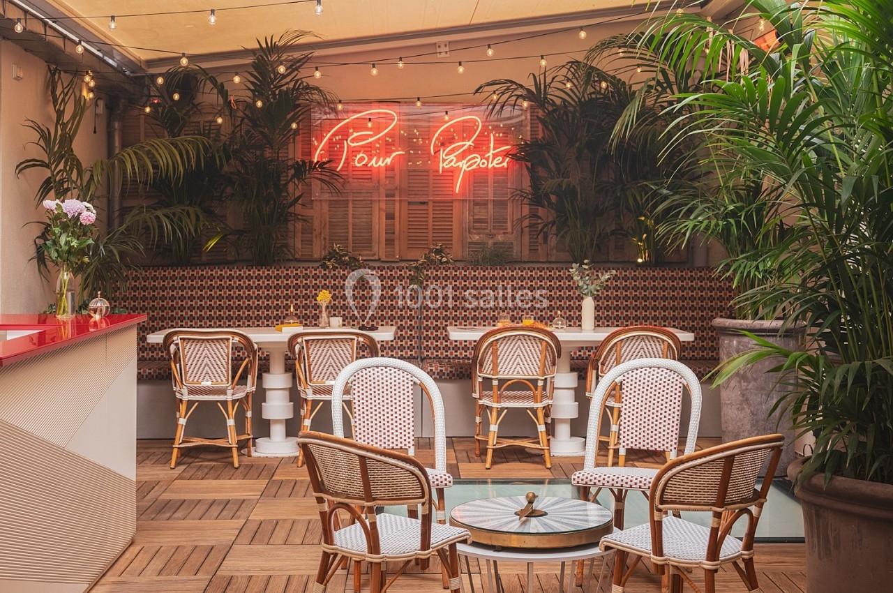 Terrasse couverte avec mobilier en rotin, plantes vertes, éclairage chaleureux et enseigne néon rouge au mur.
