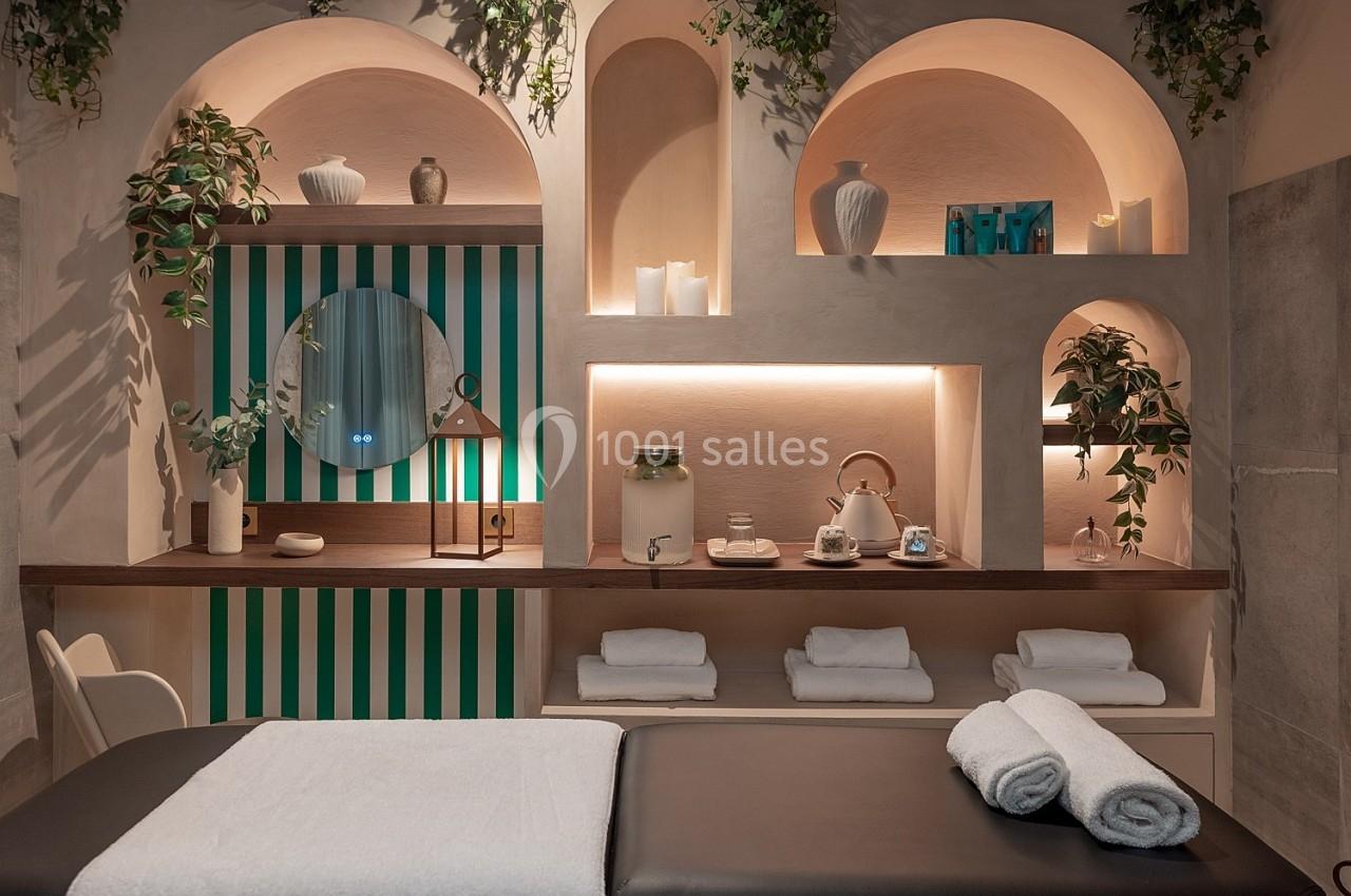 Salle de soins avec table de massage, étagères décorées de plantes, serviettes pliées et éclairage tamisé.