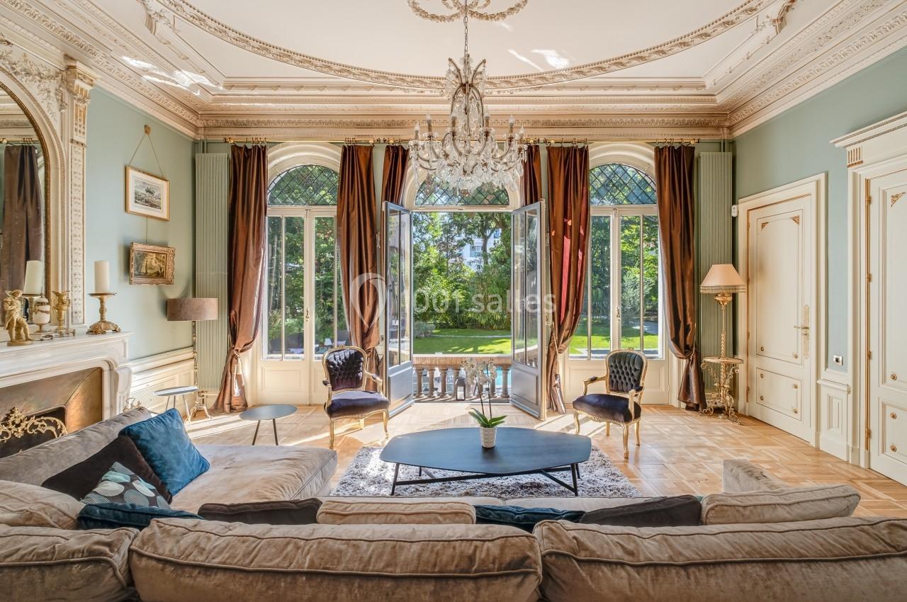 Salon élégant avec canapé beige, lustre, grandes fenêtres cintrées ouvertes sur un jardin verdoyant.