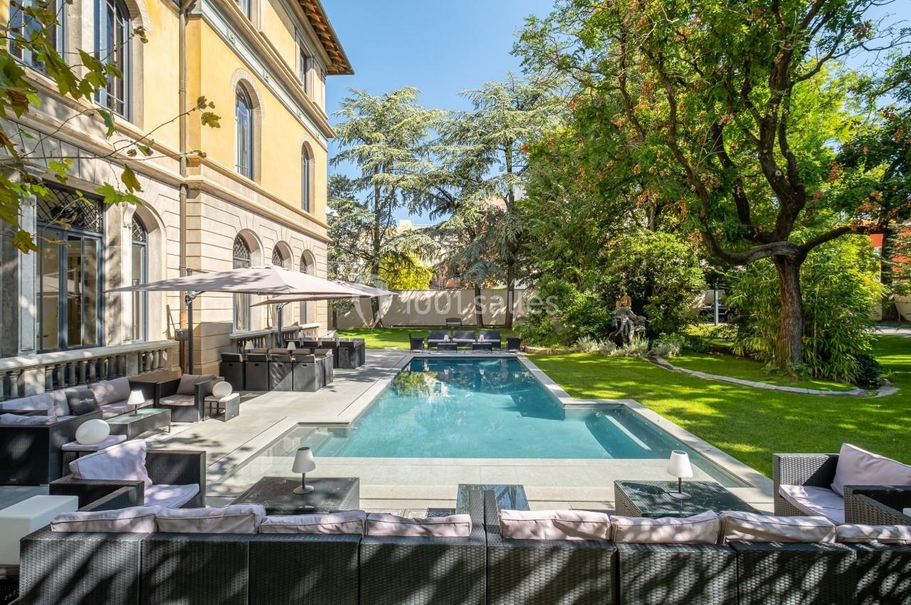 Piscine extérieure entourée de mobilier de jardin, arbres et pelouse, près d'une grande maison en pierre.