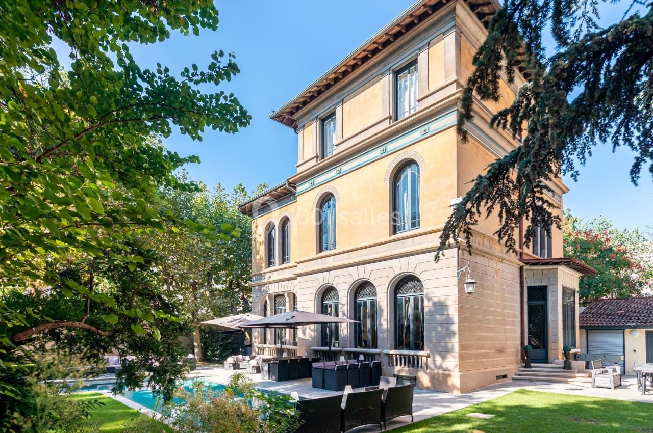 Façade d'une villa en pierre avec grandes fenêtres, entourée d'arbres, terrasse aménagée et piscine dans un jardin.
