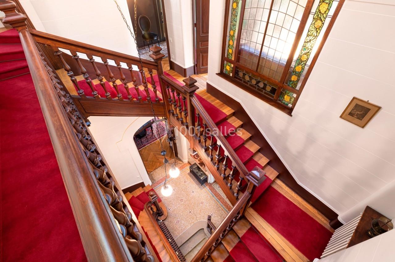 Escalier en bois avec tapis rouge, vitrail coloré sur le mur et vue plongeante sur le hall d'entrée carrelé.