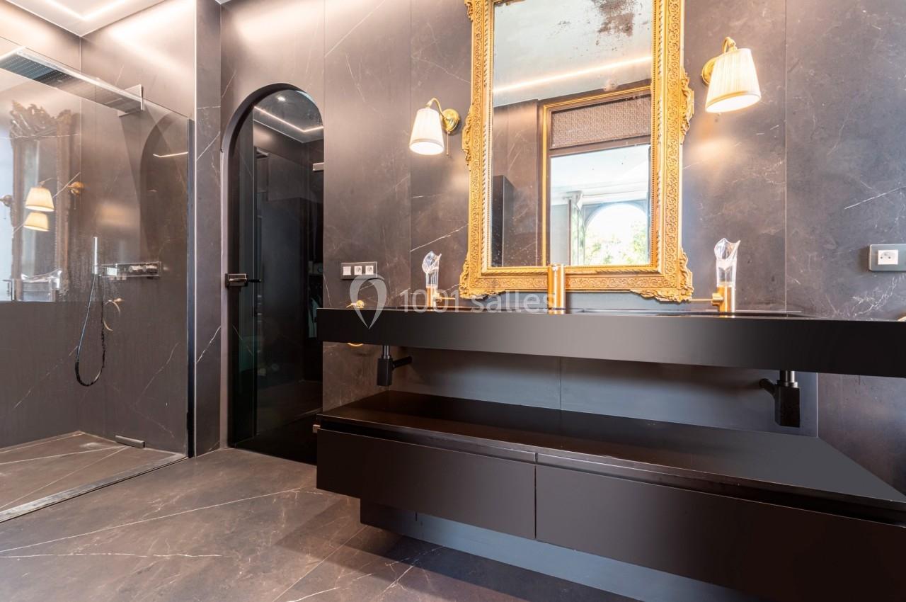 Salle de bain moderne avec murs en marbre noir, miroir doré, double vasque et douche vitrée à gauche.