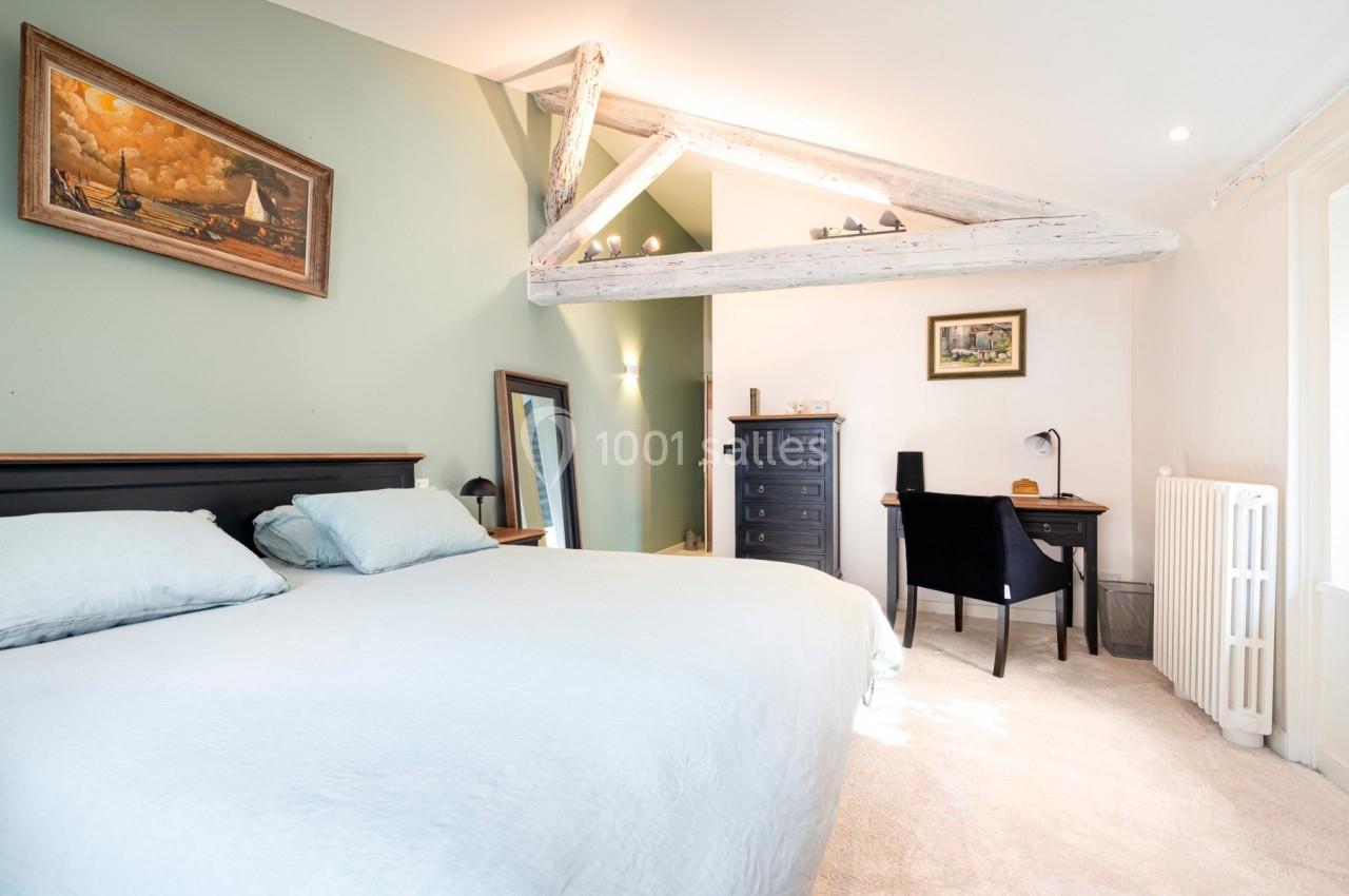 Chambre lumineuse avec lit double, poutres apparentes, bureau, commode et décorations murales dans un style chaleureux.