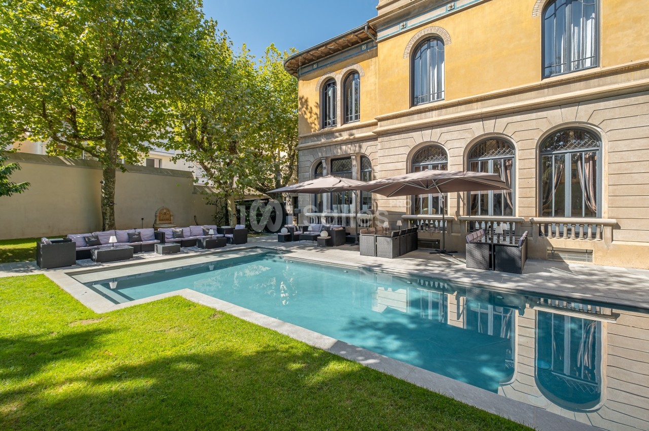 Piscine extérieure entourée de chaises longues, parasols et bâtiment ancien avec grandes fenêtres en arrière-plan.