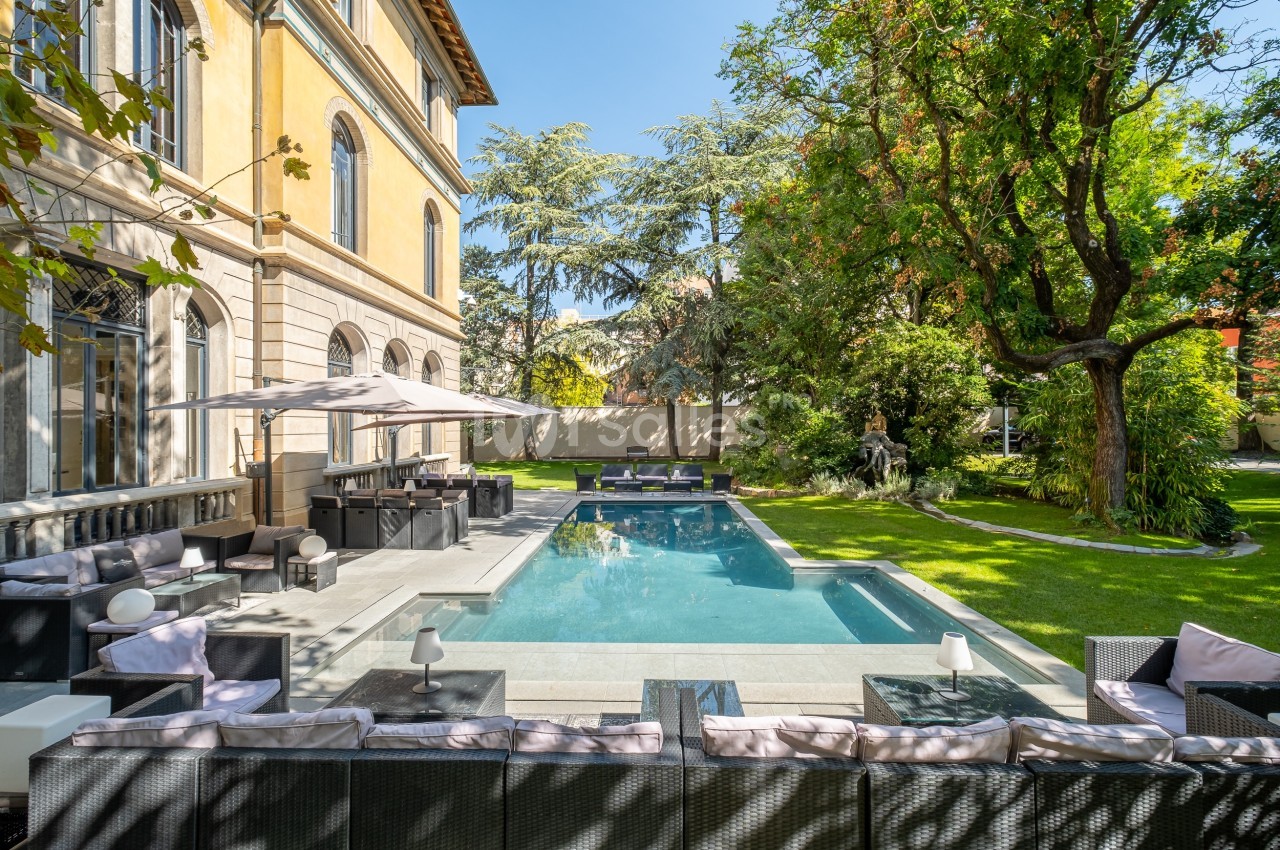 Piscine extérieure entourée de mobilier de jardin, située dans un espace verdoyant près d'une grande maison.
