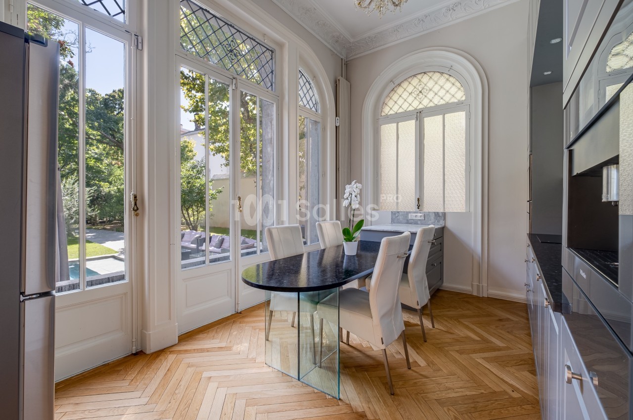 Salle à manger lumineuse avec table en verre, chaises blanches, parquet en bois et grandes fenêtres donnant sur un jardin.