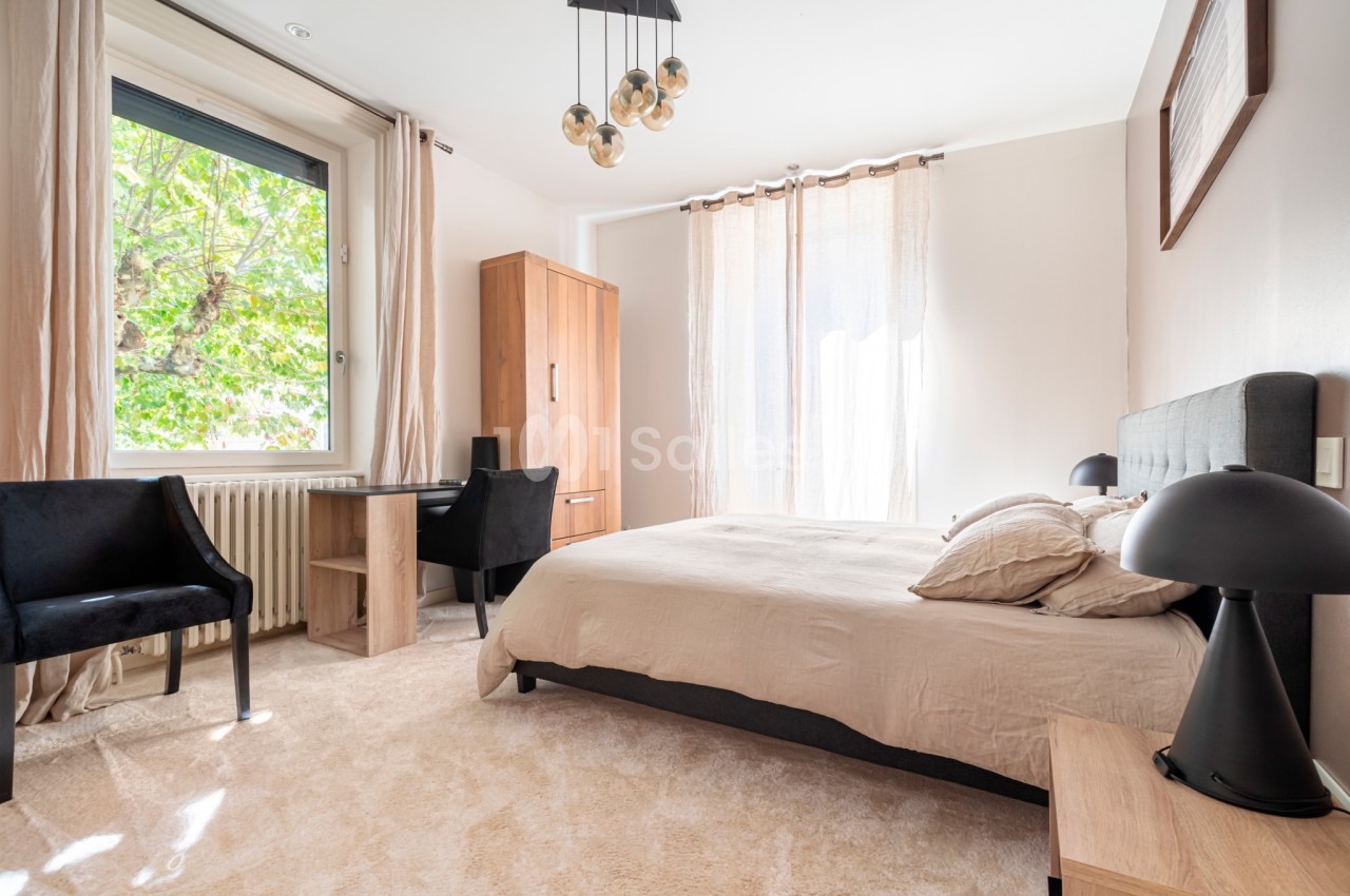 Chambre lumineuse avec lit double, bureau, deux chaises noires et grandes fenêtres donnant sur un arbre.