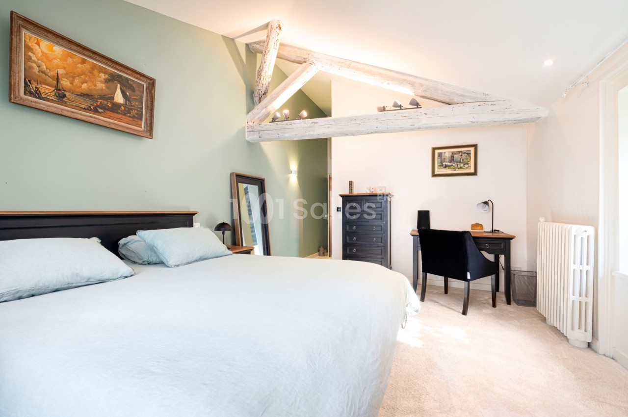 Chambre lumineuse avec lit double, bureau, poutres apparentes et murs verts, décorée avec des tableaux.