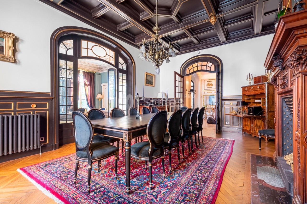 Salle à manger élégante avec table en bois, chaises noires, tapis rouge et plafond à caissons sombres.